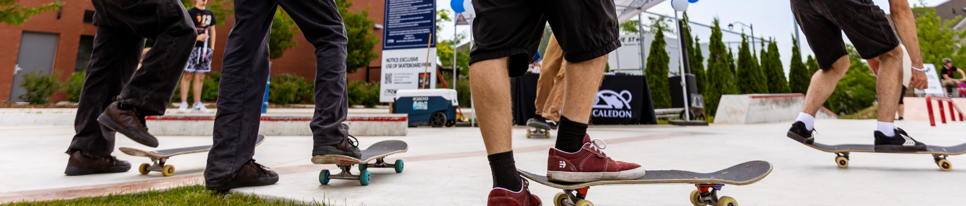 Close-up of several skateboarders at a community skatepark, with legs in motion as they ride and perform tricks on a smooth concrete surface near a Caledon event tent.