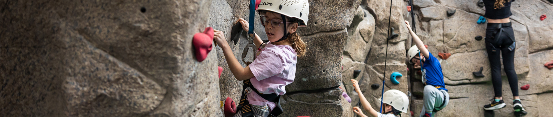Children and youth climb an indoor rock climbing wall with helmets and harnesses, participating in a supervised activity.